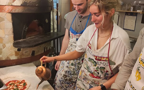 Pizza-making class participants in Naples adding oil to a pizza near a wood-fired oven.