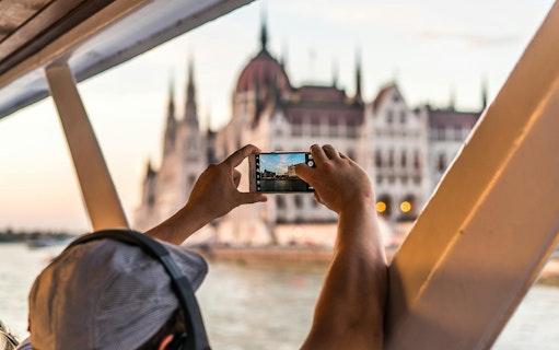 Tourist photographing Budapest Parliament from daytime hop-on hop-off cruise.