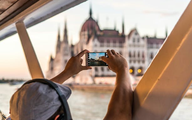 Tourist photographing Budapest Parliament from daytime hop-on hop-off cruise.