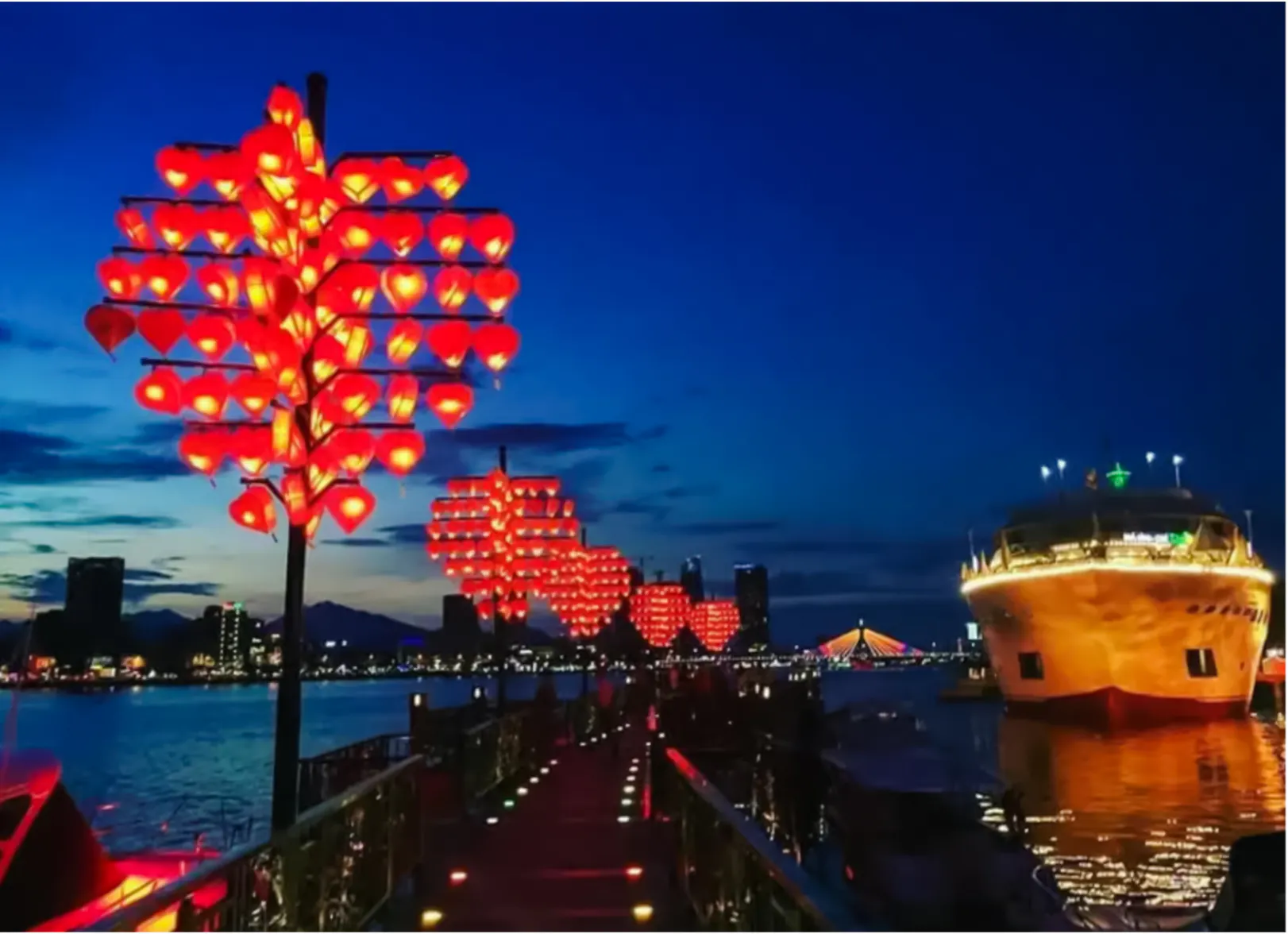 Lantern-lit walkway along Da Nang river with illuminated bridge and boat at night.