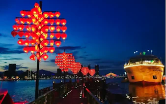 Lantern-lit walkway along Da Nang river with illuminated bridge and boat at night.