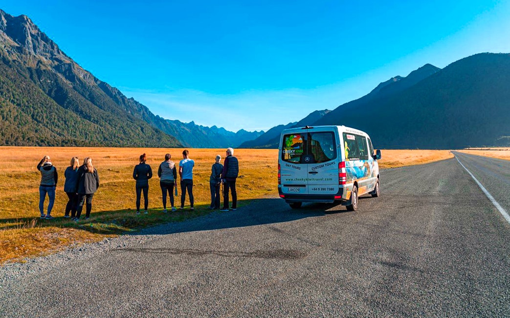 Tour group viewing Milford Sound landscape from roadside with tour van, New Zealand.