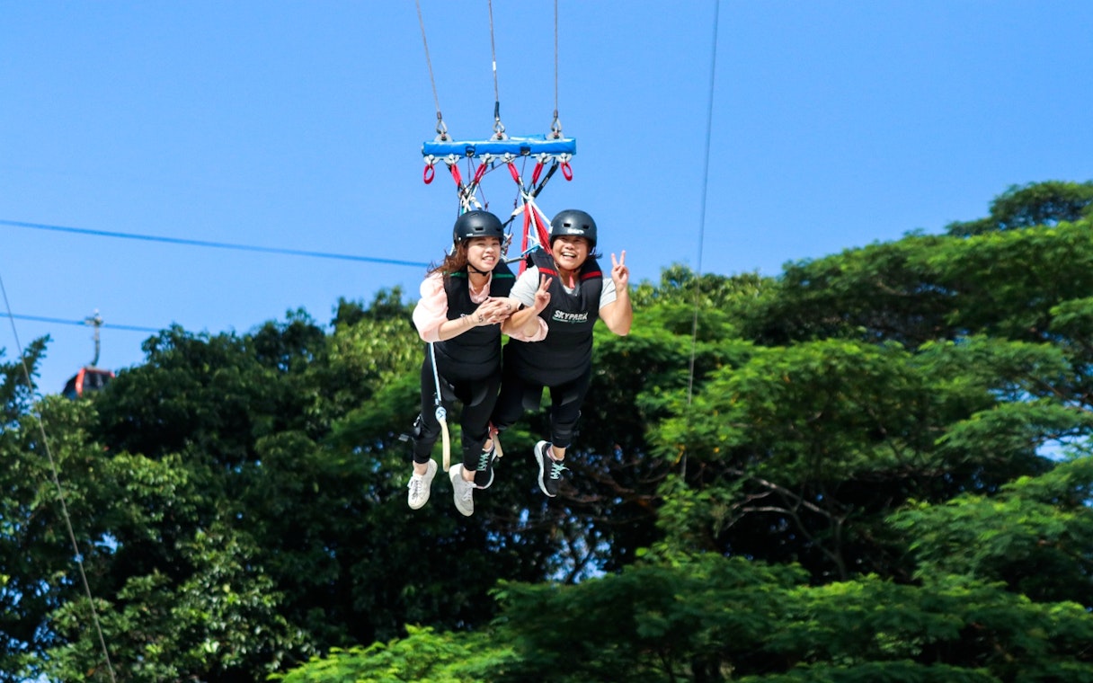 Two people tandem swinging at Skypark Sentosa by AJ Hackett, surrounded by trees.