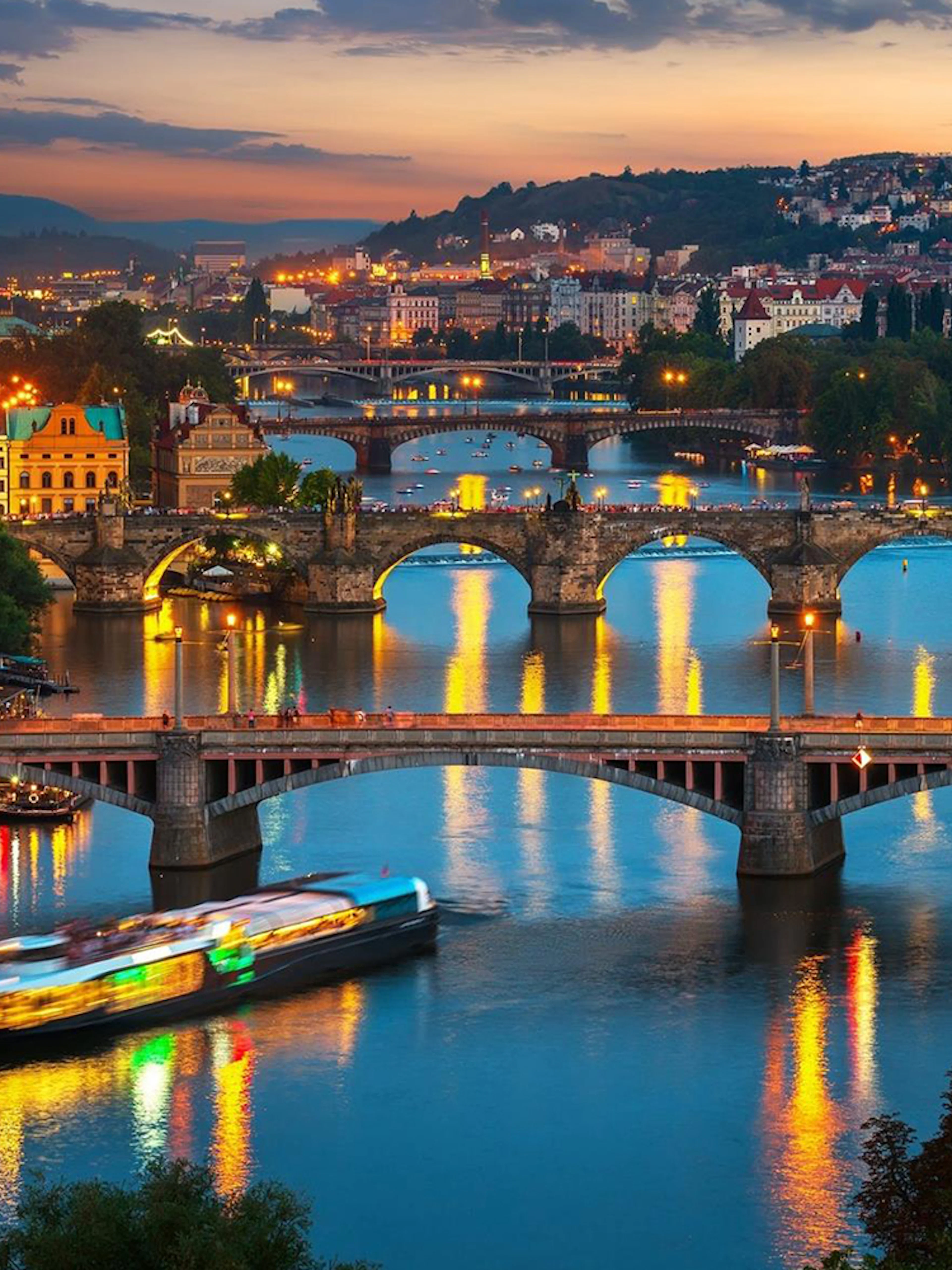 Prague river view with illuminated bridges and open-top glass boat at night.