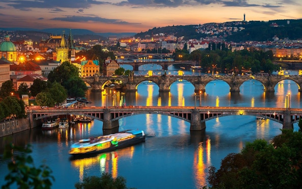 Prague river view with illuminated bridges and open-top glass boat at night.