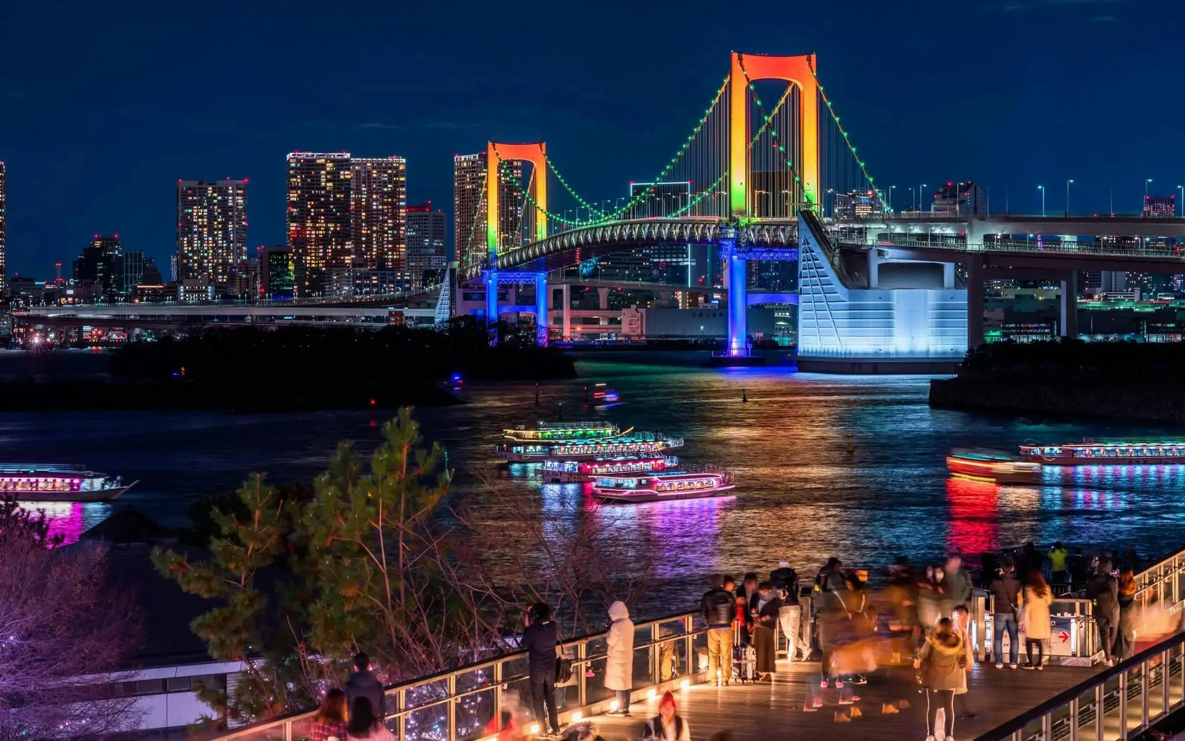 Tokyo Rainbow Bridge illuminated at night with colorful boats on the water, viewed from Daikoku PA.