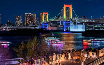 Tokyo Rainbow Bridge illuminated at night with colorful boats on the water, viewed from Daikoku PA.