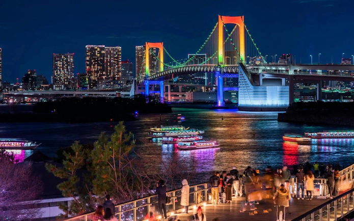 Tokyo Rainbow Bridge illuminated at night with colorful boats on the water, viewed from Daikoku PA.