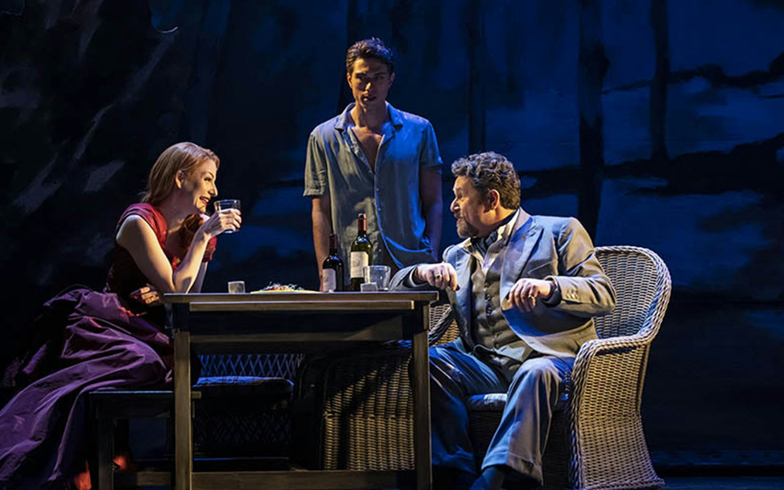 Three actors perform a scene from "Aspects of Love" at a table with wine bottles.