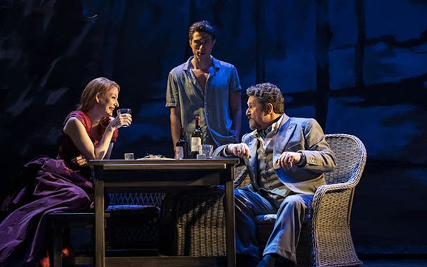 Three actors perform a scene from "Aspects of Love" at a table with wine bottles.