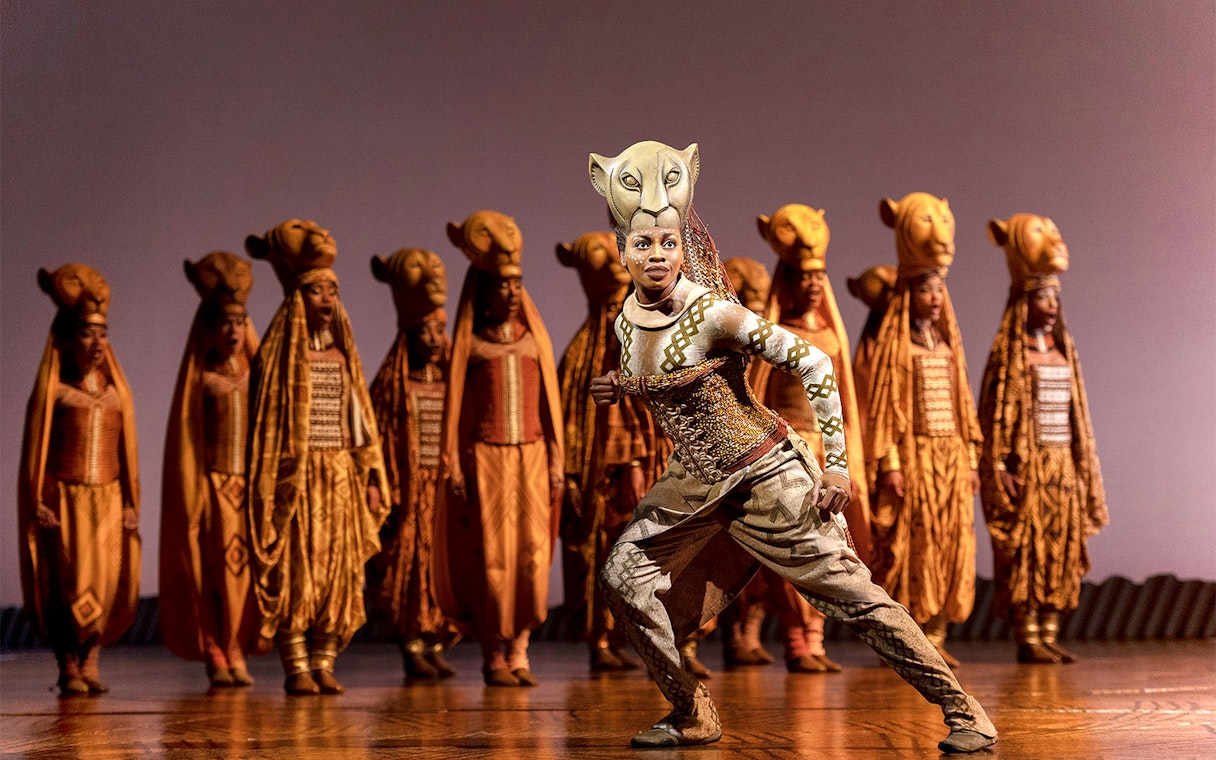 Performer in The Lion King musical with ensemble in lion costumes on stage.