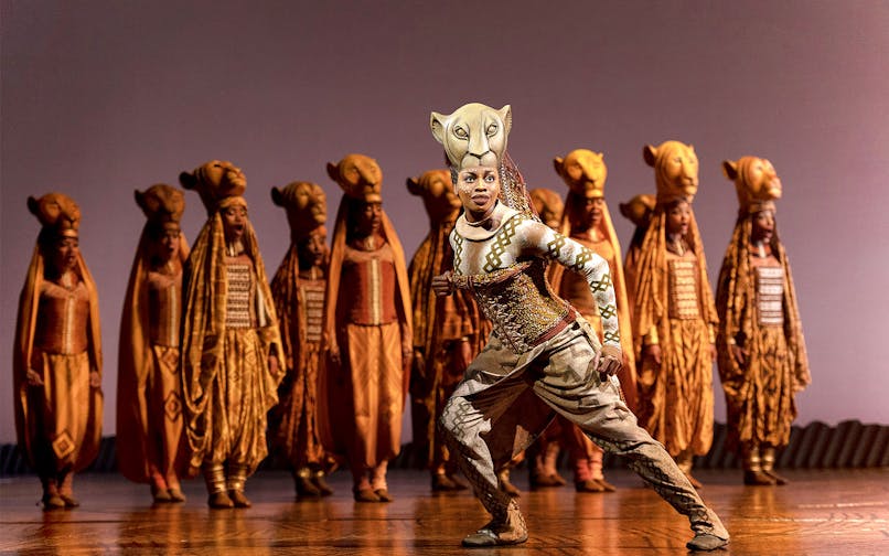 Performer in The Lion King musical with ensemble in lion costumes on stage.