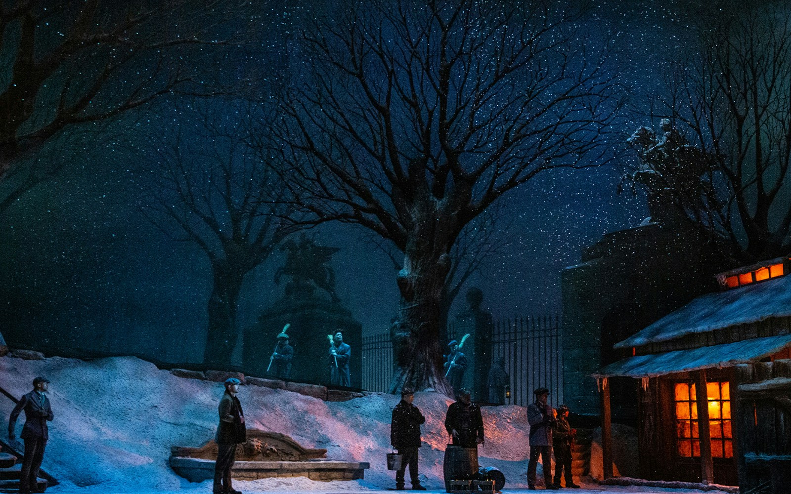 Outdoor winter scene from La Boheme with people gathered near a snow-covered building.
