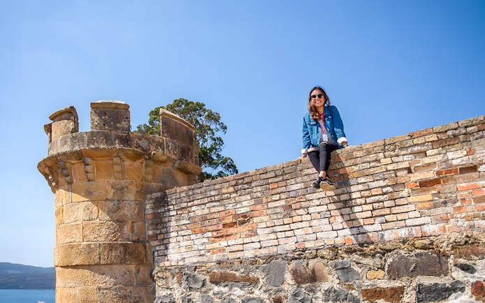Person sitting on historic brick wall at Port Arthur, Tasman Peninsula, with tower and sea view.
