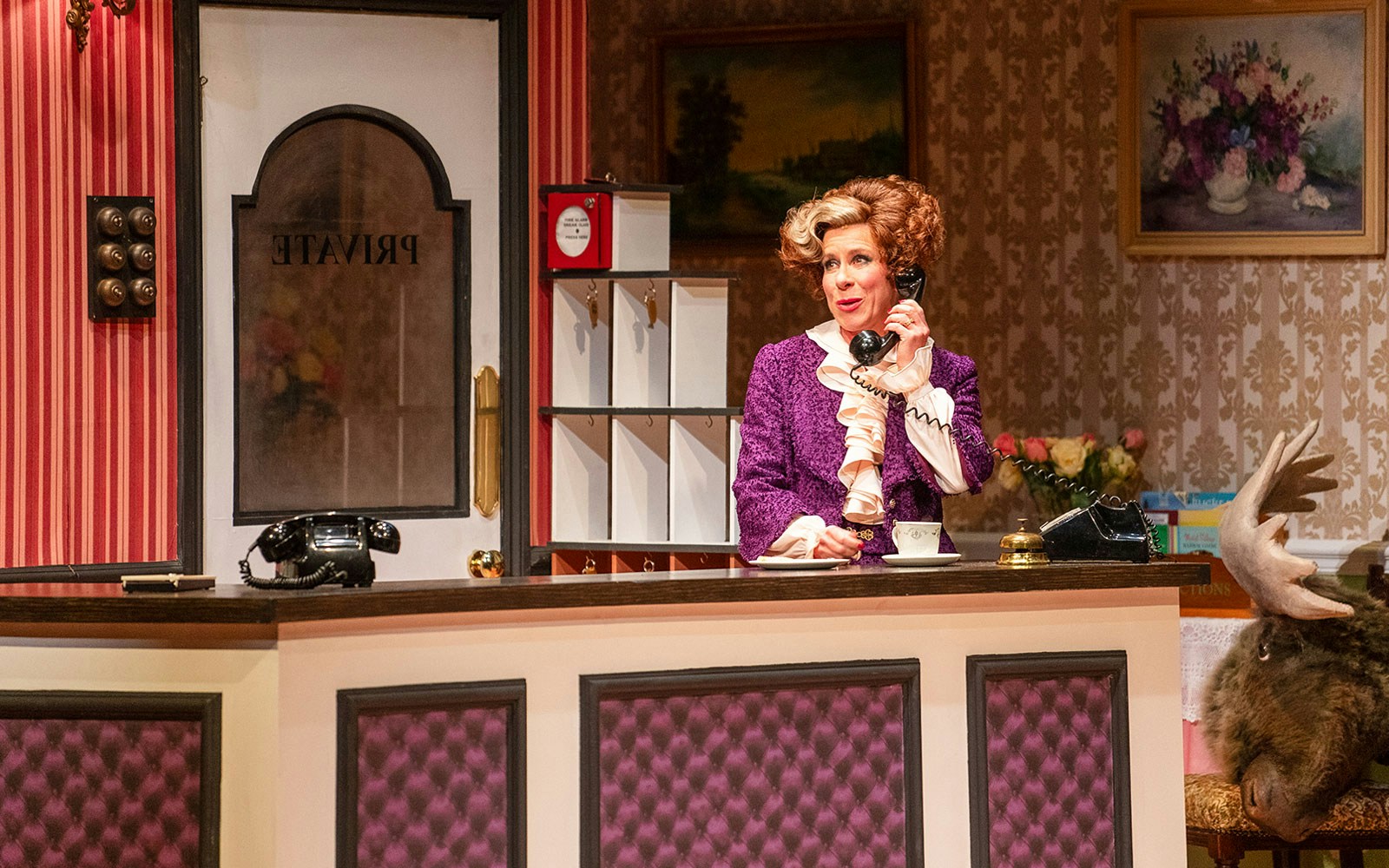 Receptionist in vintage hotel setting on phone, Fawlty Towers theme.