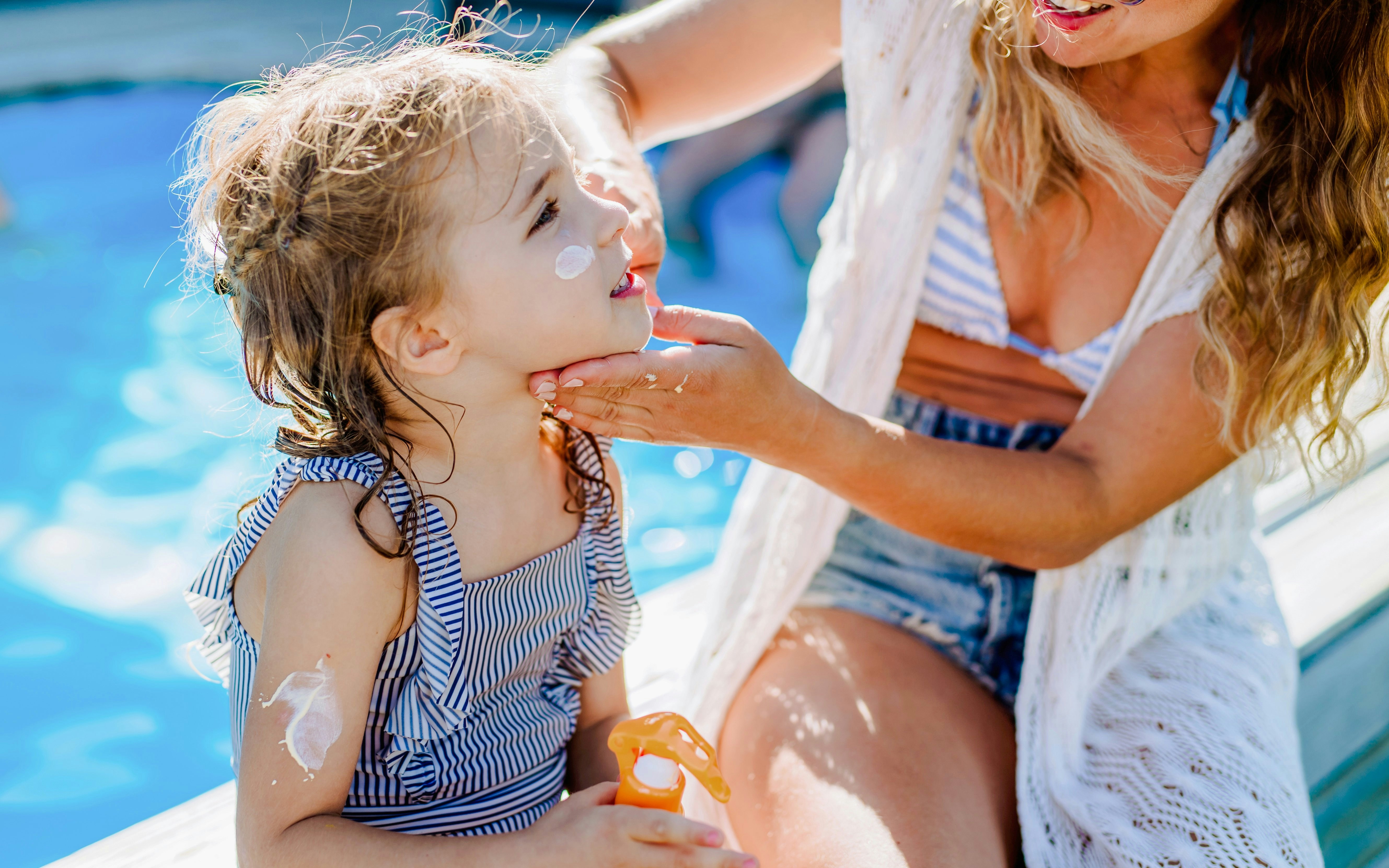 Mother applying sunscreen to daughter by a pool.