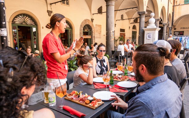 Tour guide speaking to visitors having lunch in Pisa outdoor cafe.