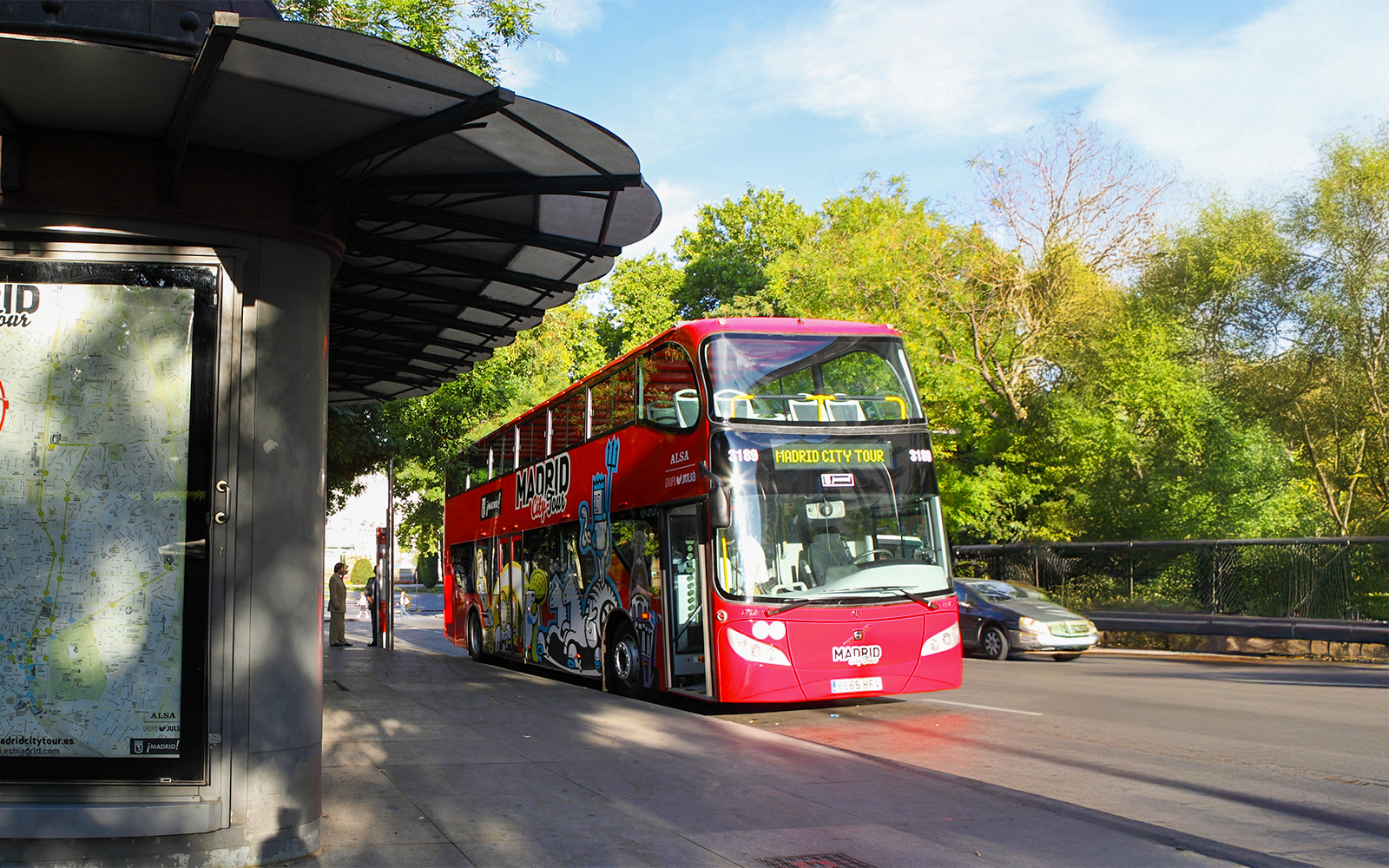 Red double-decker bus for Madrid City Tour at a bus stop with trees in the background.