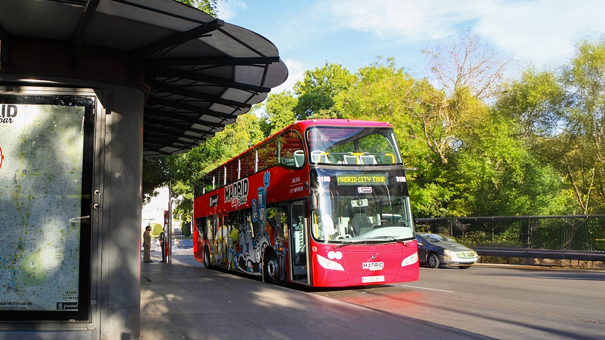 Red double-decker bus for Madrid City Tour at a bus stop with trees in the background.