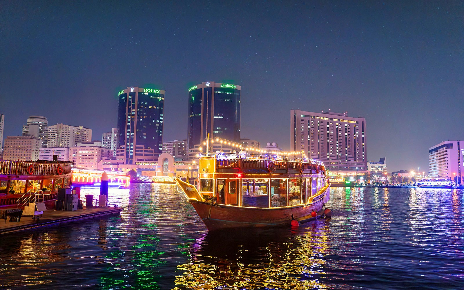 Dhow cruise with lights on Dubai Creek at night, city skyline in background.