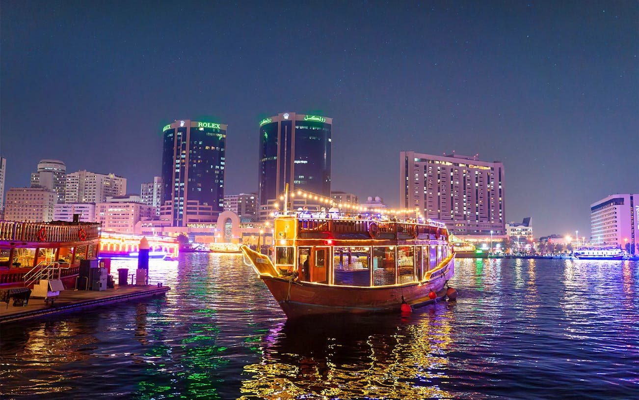 Dhow cruise with lights on Dubai Creek at night, city skyline in background.