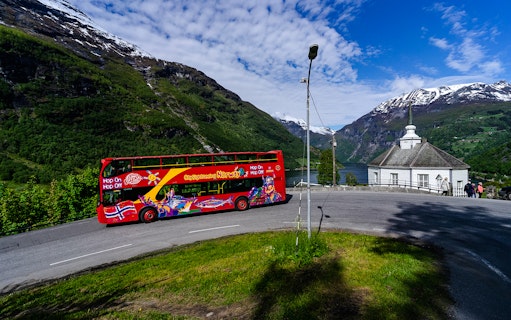Red double-decker bus on Geiranger Hop-on Hop-off tour with fjord and mountains in background.