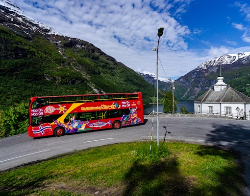 Red double-decker bus on Geiranger Hop-on Hop-off tour with fjord and mountains in background.