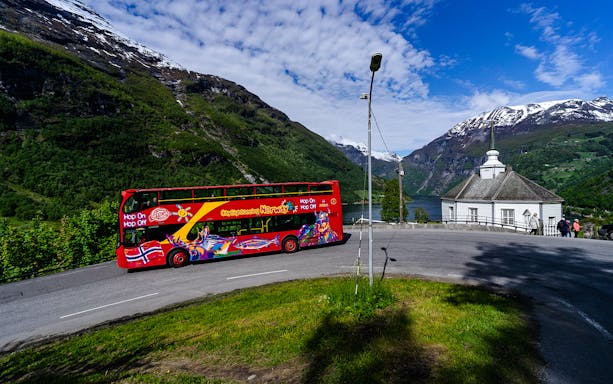 Red double-decker bus on Geiranger Hop-on Hop-off tour with fjord and mountains in background.