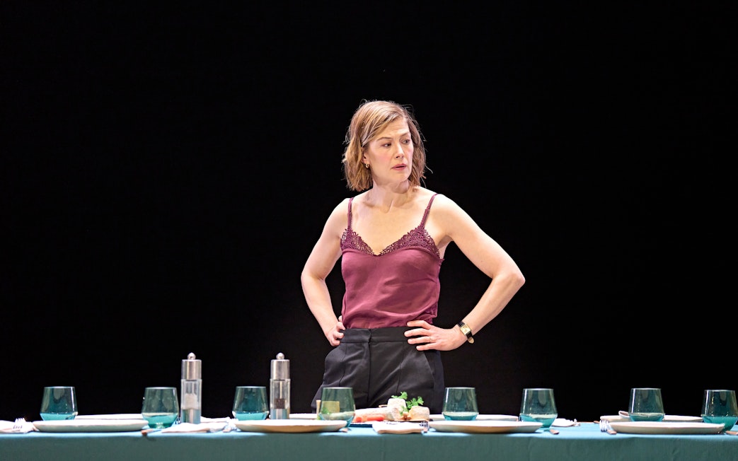 Woman standing behind a set dining table in a theatrical scene.