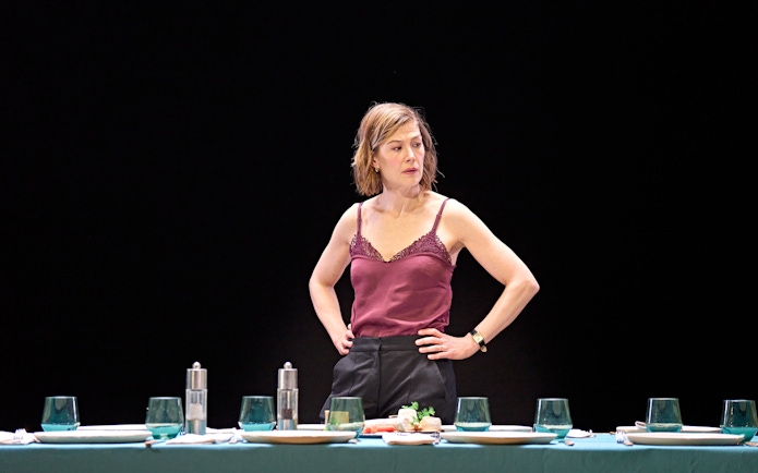 Woman standing behind a set dining table in a theatrical scene.