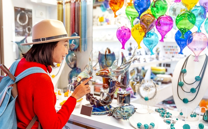 Tourist exploring glass art at Murano glass factory, colorful glass balloons and sculptures displayed.