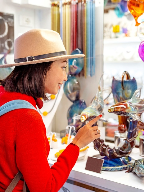 Tourist exploring glass art at Murano glass factory, colorful glass balloons and sculptures displayed.