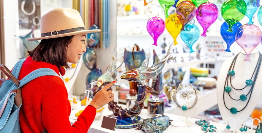 Tourist exploring glass art at Murano glass factory, colorful glass balloons and sculptures displayed.