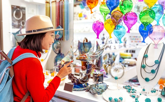 Tourist exploring glass art at Murano glass factory, colorful glass balloons and sculptures displayed.