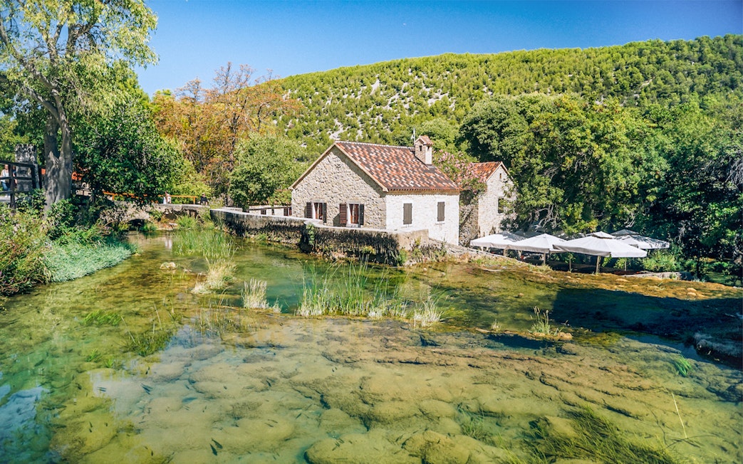 Stone house by a clear stream in Krka National Park, Croatia, surrounded by lush greenery.