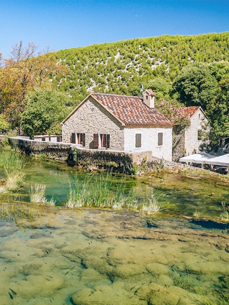 Stone house by a clear stream in Krka National Park, Croatia, surrounded by lush greenery.