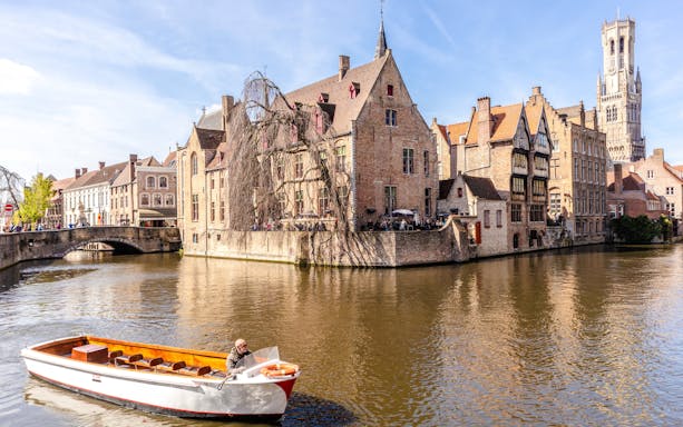 Boat on canal with historic buildings and Belfry tower in Bruges, Belgium.