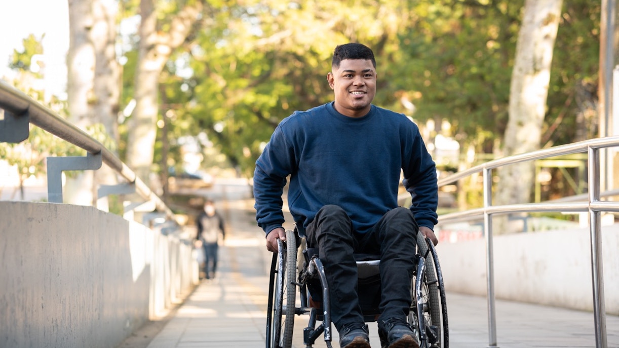 Man using a wheelchair on a sunny pathway with trees in the background.