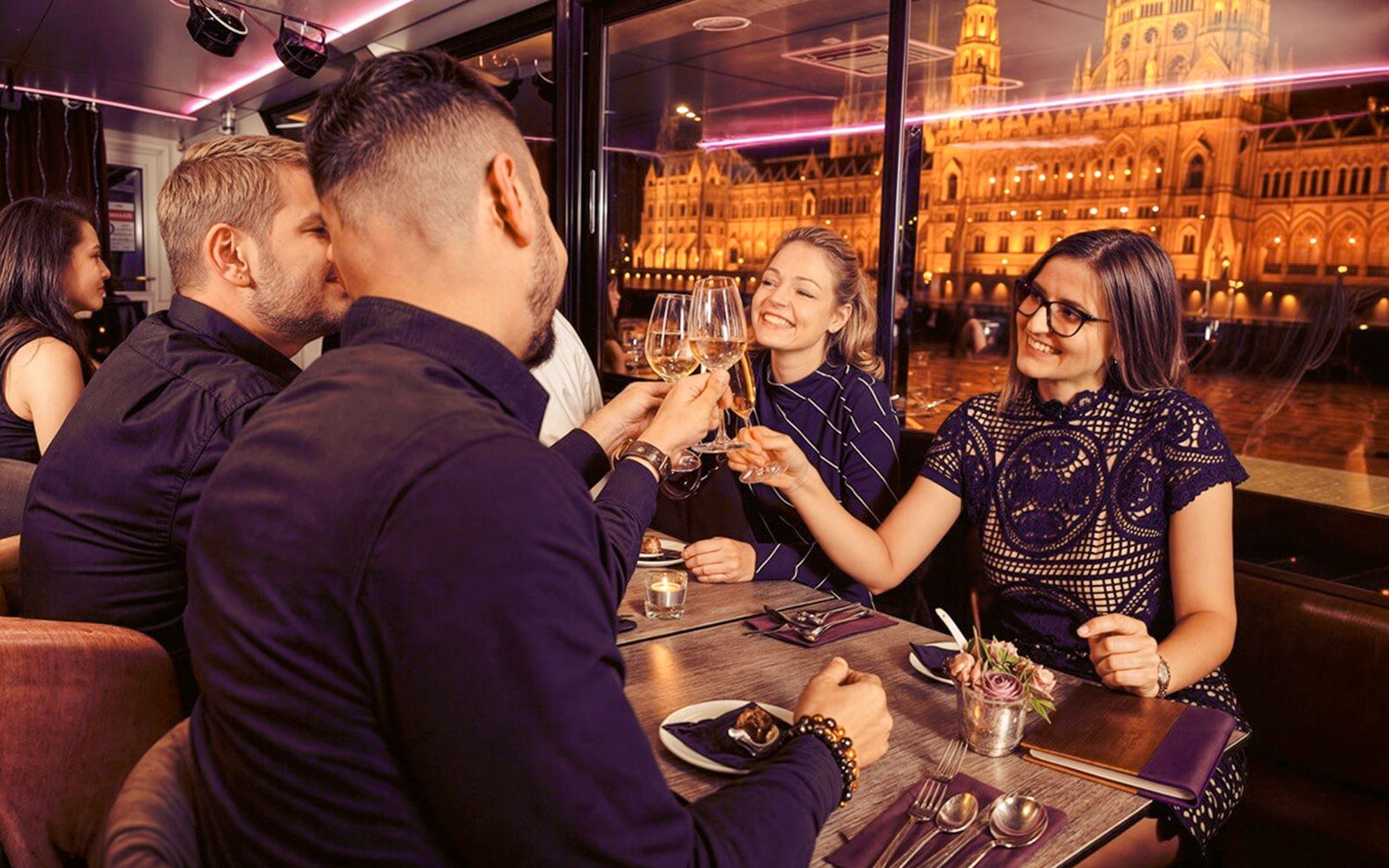 Guests toasting drinks on Budapest Dinner Cruise with Folklore Show, Hungarian Parliament lit up outside.