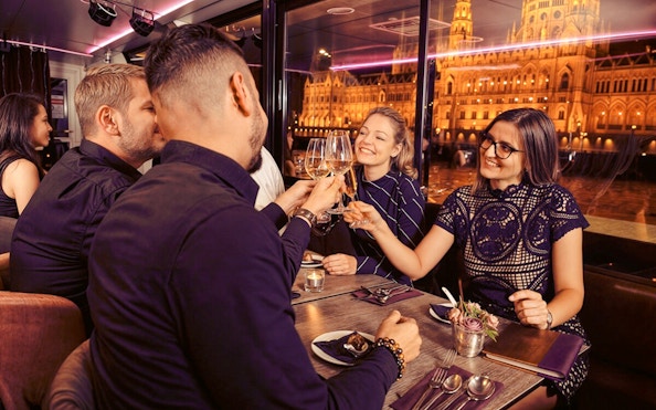 Guests toasting drinks on Budapest Dinner Cruise with Folklore Show, Hungarian Parliament lit up outside.