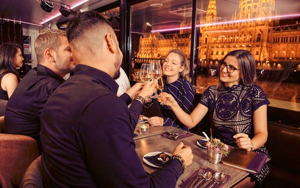 Guests toasting drinks on Budapest Dinner Cruise with Folklore Show, Hungarian Parliament lit up outside.