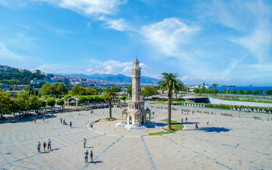Clock tower in Konak Square, Izmir city center, with people walking and sea view in the background.