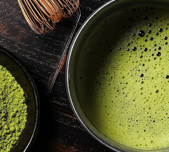 Matcha tea in a bowl with bamboo whisk and powder on a dark wooden surface.