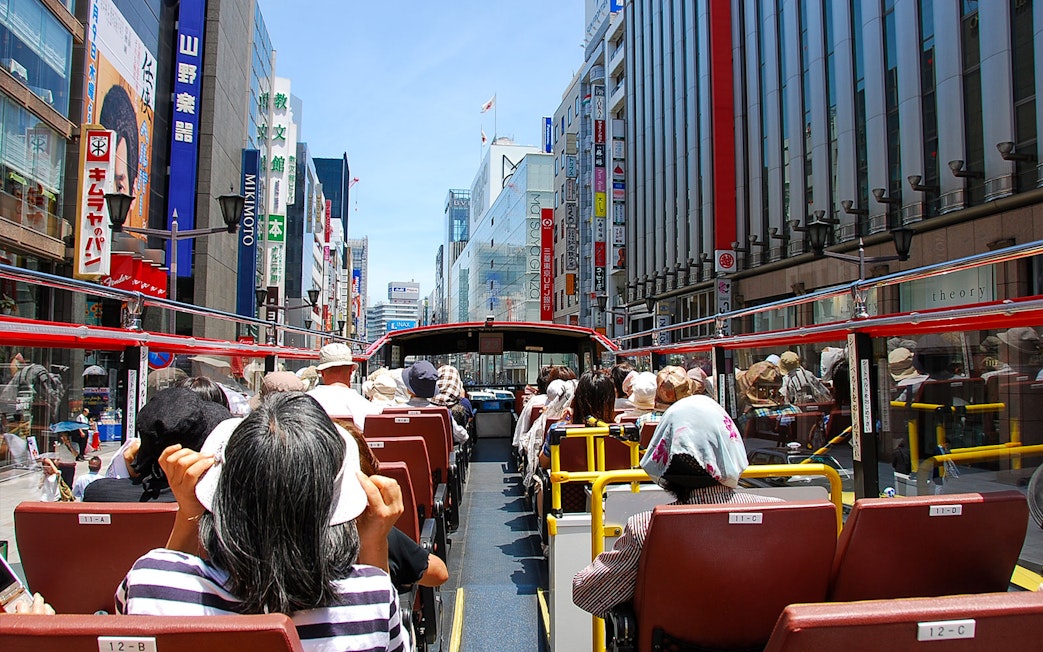 Tourists on a Sky Hop Bus tour in Tokyo, Japan, viewing city buildings and signs.