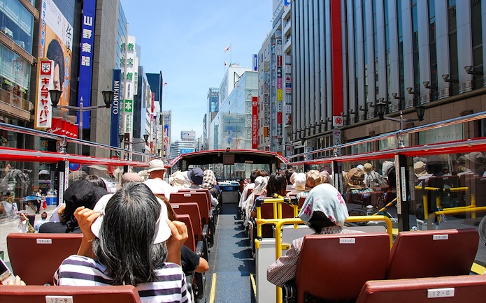 Tourists on a Sky Hop Bus tour in Tokyo, Japan, viewing city buildings and signs.