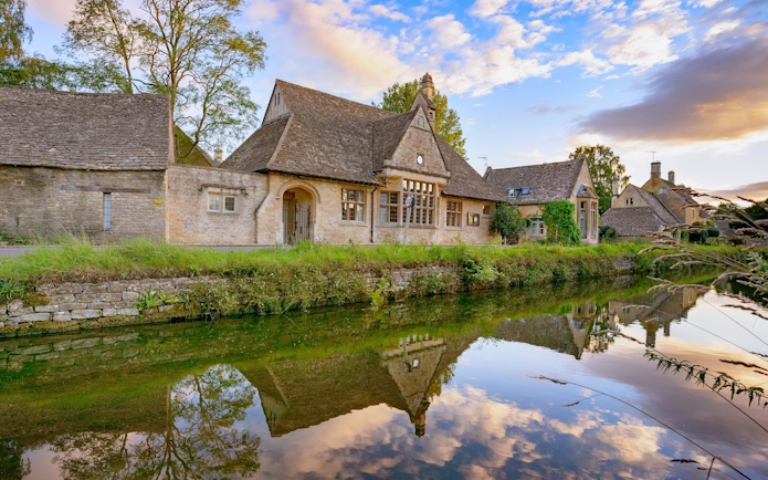 Stone cottages reflected in a tranquil river in the Cotswolds, England.