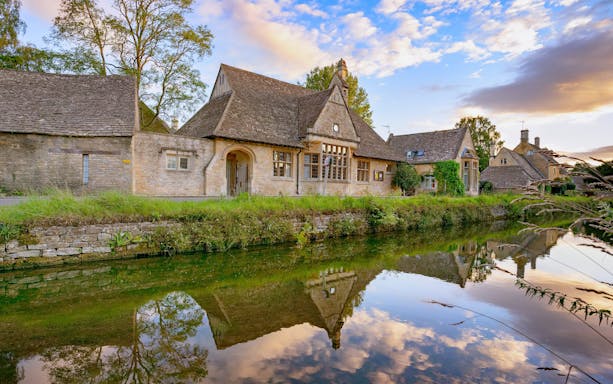 Stone cottages reflected in a tranquil river in the Cotswolds, England.