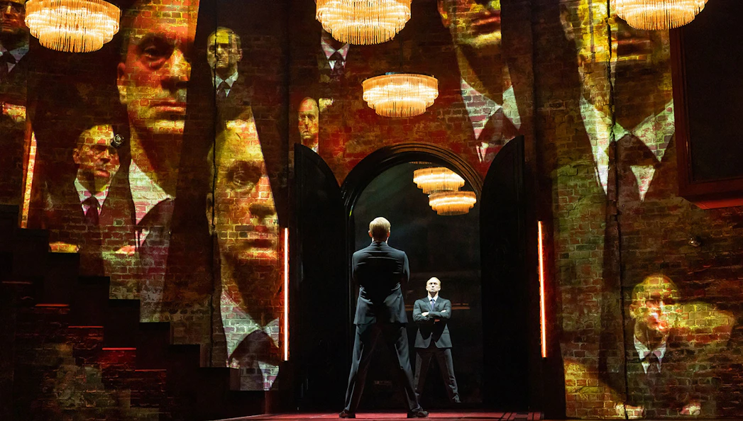 Two men in suits stand in a dimly lit room with multiple reflections on brick walls.