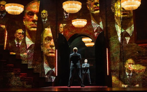 Two men in suits stand in a dimly lit room with multiple reflections on brick walls.