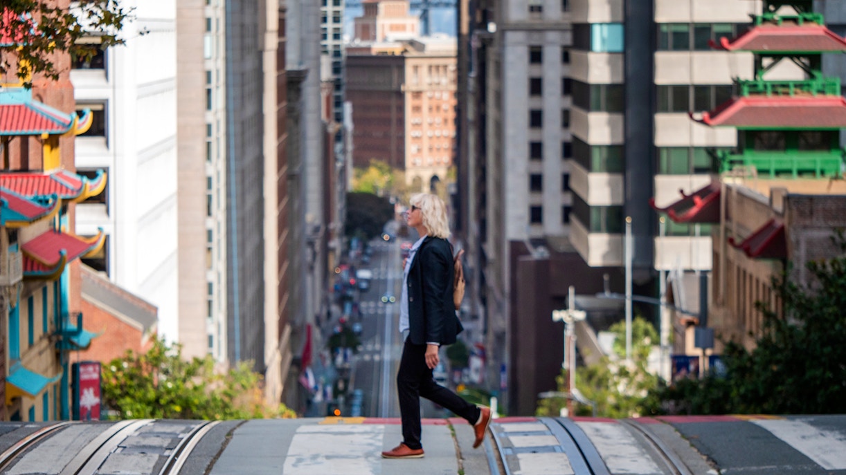 Woman walking on a hilly street in San Francisco with cityscape in background.